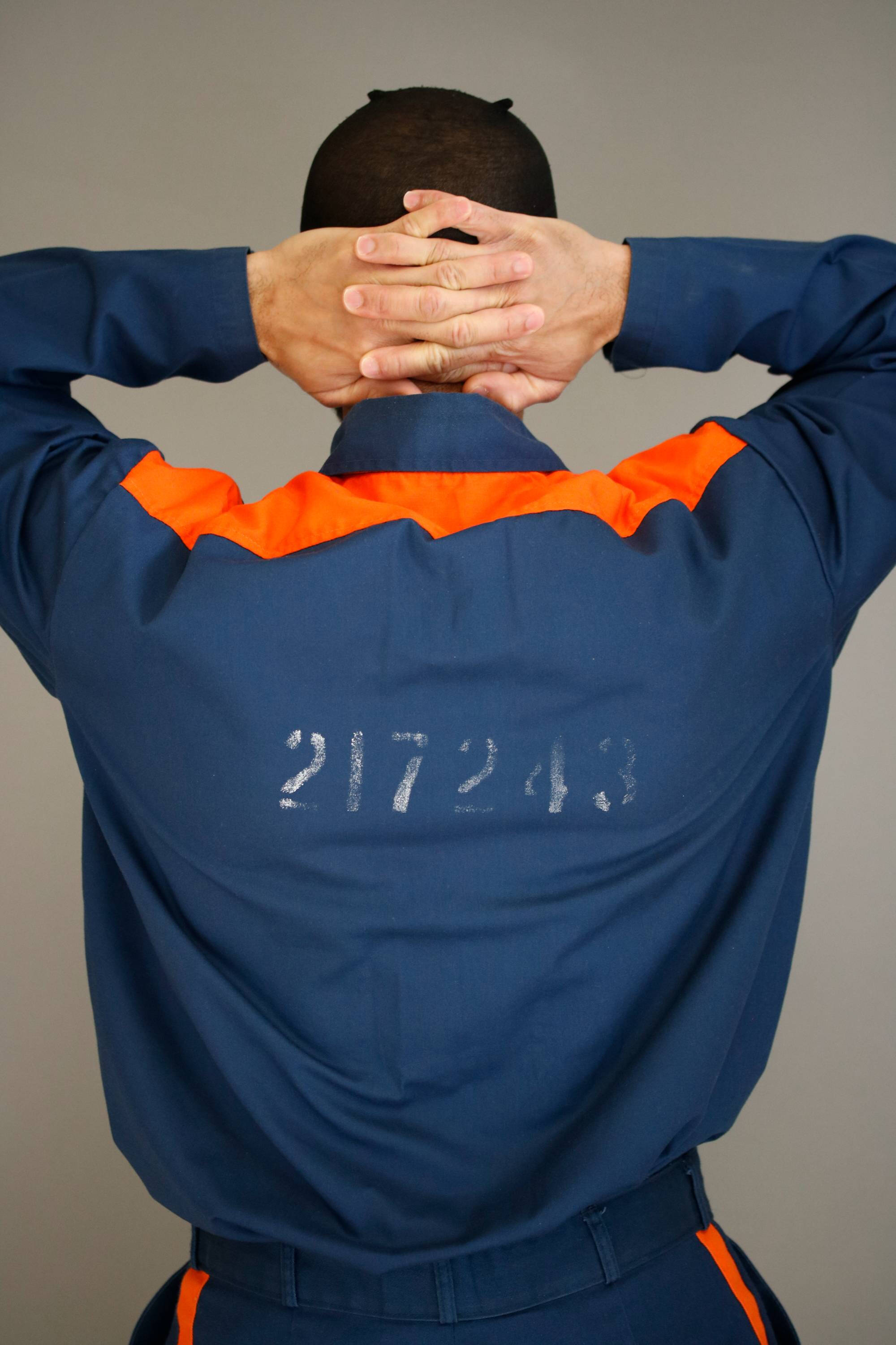 Photograph of a person with their hands clasped behind their head from behind. They wear a blue prison uniform.